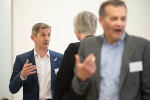Three people are engaged in conversation in a bright room. The man on the left in a blue suit is speaking animatedly to a woman facing away. The man on the right, in a gray jacket, appears to be talking or reacting to something. Captured by the UK's best event photographer, all three wear name tags.