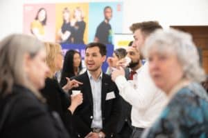 A group of people engaged in conversation at a social event. The setting appears to be indoors with a colorful, blurred poster in the background. Some individuals are holding drinks, while others are listening and talking. Captured by the UK's best event photographer, the atmosphere seems friendly and casual.