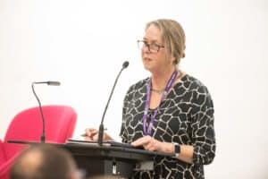 A person wearing glasses and a black and white patterned blouse stands at a podium speaking into a microphone. Holding a pen, they have a lanyard around their neck. In the background, captured perfectly by the UK's best event photographer, there's a red chair.