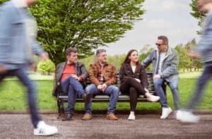 William and the Eternals casually sit on a bench in a park on a sunny day, engaged in conversation. In this serene Manchester setting, the background shows grassy areas and trees. In the foreground, two blurred figures walk past, adding a sense of motion reminiscent of candid comedy photography. This is the cover of their single.