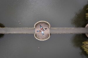 Aerial view of five members of a band backs with arms outstretched in a circular area of a bridge over a body of water, reminiscent of an artistic scene one might find in music and comedy photography from Oldham. Several ducks swim nearby. The bridge spans across the water with the circular area as a scenic viewpoint.