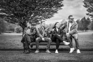 Black and white image of four people sitting on a park bench in Oldham. They all wear casual attire, including jackets and jeans. One person is looking left, another appears relaxed, a woman is smiling and talking, and a man on the right is adjusting his sunglasses. Trees and grass form the background.