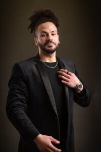 A man with dark, curly hair tied up, a beard, and a mustache poses against a dark background. He is wearing a black blazer, a black shirt, a necklace, and a wristwatch. His right hand touches his chest while his left hand rests by his side—a perfect capture by the head shot photographer in Manchester.
