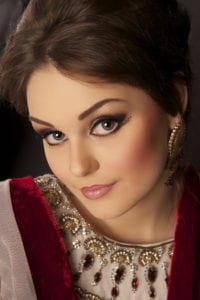 A woman with dark hair and pronounced makeup gazes at the camera, captured expertly by an Oldham photographer. She is wearing a red and white dress adorned with intricate beadwork and gemstone decorations. Her expression is calm and confident, accentuated by an elegant earring in one ear.