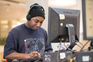 A person wearing a black beanie and headphones is focused on using a computer in front of them, dressed in a dark t-shirt with a large "SC" logo on the front. The light indoor setting, with its blurred details, resembles the meticulous style of an Oldham photographer capturing the moment.