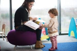 A woman wearing glasses sits on a purple stool, reading a large picture book to a small child standing beside her. The child is attentively looking at the book. Captured by an Oldham photographer, they are in a well-lit room with large windows and vibrant children's furniture.