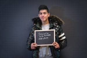 A person with dark hair, wearing a shiny black puffer jacket with a fur-lined hood, is holding a small chalkboard. The handwritten message reads, "Socialise with friends and chill." Captured by an Oldham photographer, the scene's background is plain and dark.
