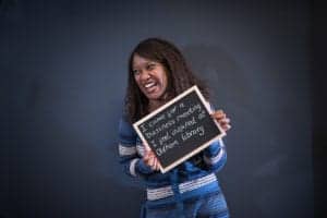 A woman with long hair smiles while holding a small chalkboard. The chalkboard reads, "I came for a business meeting... I feel inspired at Oldham Library." She is dressed in a blue and white striped outfit and stands against a plain dark background, captured perfectly by an Oldham photographer.