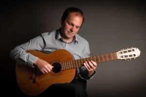 Oldham musician Mike Mellor in a grey shirt plays a classical guitar against a dark background, embodying the calm and focused mood often seen in music photography. They look down at the fretboard, with their right hand plucking the strings and their left hand pressing down on the neck.