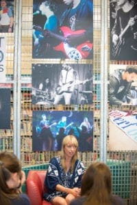 A woman with blonde hair wearing a blue dress with white patterns sits in a red chair, facing a small group of people. Behind her, large photographs by an Oldham photographer of musicians playing guitars are displayed on a wall of metal mesh. The setting appears to be an informal talk or seminar.