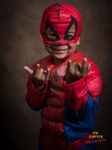 A young child dressed in a Spider-Man costume poses with a big smile, holding his hands out in web-shooting gestures. The background is a neutral brown, and the bottom right corner displays "Tim Simpson Photography" in green and red text, showcasing the vibrant talent of this Oldham photographer.