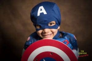 A child dressed in a Captain America costume smiles at the camera, holding a red, white, and blue Captain America shield. The background is dark brown and the name "Tim Simpson Photography" is written in the bottom right corner by renowned Oldham photographer.