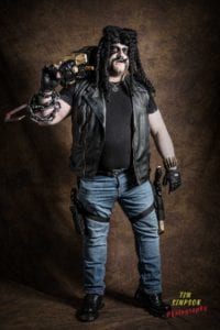 A person is dressed in a leather vest, black shirt, jeans, and black boots, holding a large weapon. They wear dark face paint, black gloves, and a chain with a hook. The textured brown backdrop enhances the dramatic scene. Text in the corner reads "Tim Simpson Photography," capturing the essence of an Oldham photographer's vision.