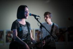 A woman with a black bob hairstyle, wearing a t-shirt and playing an electric guitar, sings into a microphone on stage. Next to her, in the background, is a man playing another guitar. Captured by an Oldham photographer, the setting exudes the energy of a live music performance.