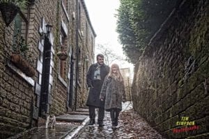 A man and woman walk closely together down a narrow, cobblestone street between two stone buildings. They are both wearing long coats and scarves. A cat sits on the edge of the path, and rain gives a glistening effect to the scene. "Tim Simpson Photography, Oldham Photographer" is in the corner.