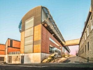 A modern building with a mix of glass, wood, and metal elements. An elevated walkway connects it to an older, stone structure on the right. The area around the buildings is clean and well-maintained, with clear skies in the background – perfect for an Oldham photographer to capture its charm.