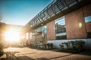 The image, captured by an Oldham photographer, showcases a modern building made of glass and brick with a distinctive tube-like structure on top. The sun shines brightly from the left side, casting a warm glow over the scene. Lush greenery and a paved walkway lead to the entrance.