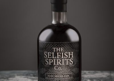 A dark glass bottle of "The Selfish Spirits Co. Dark Spiced Rum" with a black label and wax-sealed cap, displayed against a dark, textured background. This exquisite 70cl bottle with an alcohol content of 40% ABV showcases the artistry of product photography Manchester is renowned for.