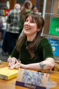 A woman with long brown hair sits at a table, smiling and holding a pen. She wears a green velvet top and has a tattoo on her forearm. In front of her are two books, one with a yellow cover and the other titled "Fantasy." With the eye of the UK’s best event photographer, shelves brimming with books frame the scene.