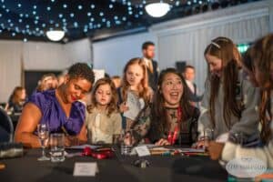 A diverse group of women and children sit around a table, engaged in a lively and animated discussion. They are surrounded by art supplies, papers, and drinks. The background is decorated with string lights, creating a festive atmosphere. A watermark from the UK's best event photographer is visible.
