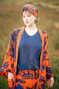 A person with light skin and light hair stands outside in a grassy area, wearing a blue shirt underneath an orange and blue patterned open robe with matching pants and headscarf. The background is blurry, focusing on the person, captured perfectly by a head shot photographer in Manchester.