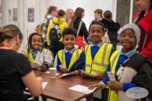 A group of four smiling children in high-visibility vests stand at a table, holding papers and cards. An adult sits on the opposite side, engaging with them. Other people are visible in the background. The image has a watermark that reads "Tim Simpson," known as the UK's best event photographer.