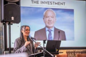 A woman in a gray blazer stands at a podium, speaking into a microphone with an open laptop in front of her. Behind her, an image captured by the UK's best event photographer showcases "THE INVESTMENT" and an older man in a suit. A large speaker is visible to the left.