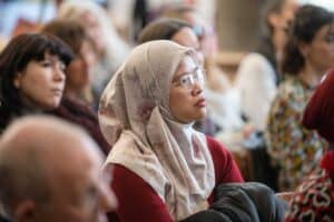 A group of people seated and attentively listening. A woman in the foreground is wearing glasses and a floral patterned hijab, with a red sweater and black jacket. Captured with precision by the UK's best event photographer, other individuals with varying expressions are visible in the background.