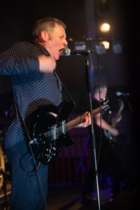 A man passionately sings into a microphone while playing an electric guitar on stage. With the UK's best event photographer capturing every moment, he wears a dark polka dot shirt and blue jeans. Behind him, another band member also plays an electric guitar. The stage is lit by warm lights, creating an energetic atmosphere.