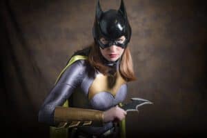 A person dressed in a Batgirl costume stands in a powerful stance against a dark, blurred background. They are wearing a black mask with bat ears, a purple and gold suit, and a yellow cape. Captured by the UK's best event photographer, they hold a bat-shaped weapon in one hand, looking determined.