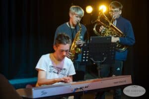 A young man in a white Adidas shirt plays a keyboard on stage. Behind him, another young man with a saxophone and another with a French horn prepare to play. A music stand with sheet music is in front of them. Stage lights glow in the background, captured perfectly by the UK's best event photographer.