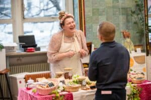 A woman with a flower headband and apron is smiling and talking to a man at a decorated market stall indoors; various food items like jars and baskets are on the table. The cheerful, inviting setting is perfectly captured by the UK's best event photographer.