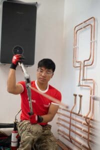 A person wearing a red shirt and camo pants works with copper pipes and a press tool in a white-walled room. Several copper pipes extend behind them along the wall with precise bends and joints, and a black rectangular device is mounted overhead—like they're being captured by the UK's best event photographer.