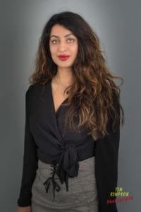 A woman with long, wavy hair, wearing a black blouse and a checkered skirt, is standing against a plain backdrop. She has red lipstick and is looking at the camera with a neutral expression. The text "Tim Simpson Photography - Oldham Photographer" is visible in the bottom right corner.