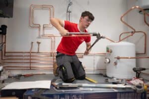A person in a red shirt and safety goggles works on copper piping in a room filled with various plumbing equipment, reminiscent of the meticulous focus of the UK's best event photographer. They are using a wrench while kneeling on the floor, completely absorbed in their task.