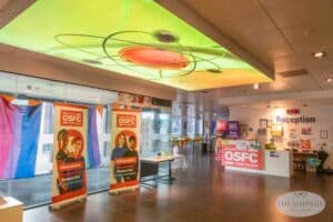 The image shows the interior of Oldham Sixth Form College, captured by the UK's best event photographer. It features a brightly lit ceiling decoration, banners with student faces and college logos, a reception desk, and various informational displays. The space is modern with colorful accents.