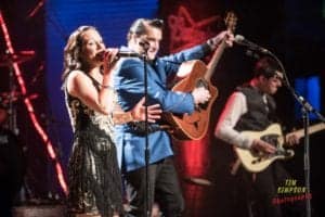 Three musicians perform on stage in a well-lit setting. Captured by an Oldham photographer, a woman sings into a microphone, wearing a black and gold dress. Two men play guitars, one in a blue blazer and the other in a dark vest. Musical equipment and lights are visible in the background.