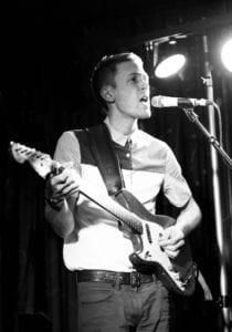 A black and white image of a male musician playing an electric guitar and singing into a microphone on stage. He is wearing a collared shirt and has a guitar strap over his shoulder. A spotlight shines on him from above, highlighting his face and guitar.