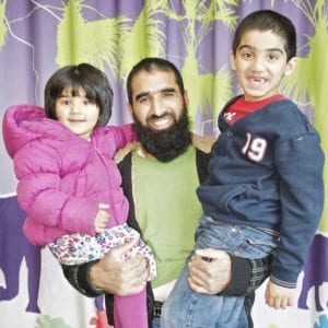 A man with a beard smiles while holding a young girl in a pink jacket and a young boy in a navy blue jacket. The children are also smiling. Captured by an Oldham photographer, the background features a colorful curtain with abstract tree and animal designs.