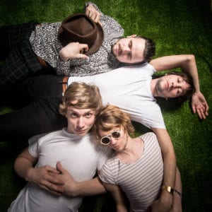 Four young men including Jake Fletcher are lying on green grass, close together. One wears a hat and patterned shirt, another in a white T-shirt with sunglasses, one in a striped shirt, and the last in a white T-shirt with a relaxed pose. Captured by an Oldham photographer, they appear to be enjoying a casual moment together.