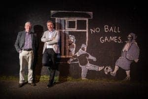 Two men stand in front of a dark wall with street art depicting three boys playing with a ball and an elderly woman leaning out of a window. The words "NO BALL GAMES" are painted next to the art. Captured by an Oldham photographer, the men are smiling, and one has his arms crossed.