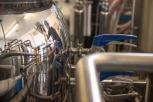 A worker in a brewery is reflected in the shiny surface of a large metal fermenter tank. They are wearing a hairnet and pouring ingredients through a funnel into the tank. The scene is filled with stainless steel brewing equipment and piping.
