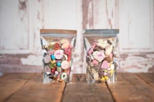 Two clear, resealable bags filled with assorted colorful candies and chocolates sit on a rustic wooden surface. Each bag has a round, pink label with the word "Lily" on it. The background features a weathered, white wooden wall.