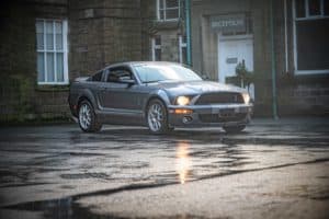 A grey Ford Mustang Shelby GT500 is parked on a wet road in front of a stone building with a "Reception" sign above the door. The scene appears to be after rainfall, with reflections visible on the wet ground.