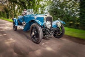 A person drives a vintage blue open-top car on a country road lined with green trees. The car features prominent round headlights, a sleek chrome grille, and black wire-spoke wheels. The motion blur effect emphasizes the car's speed and movement.