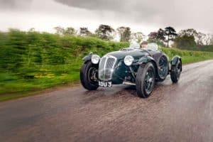 A vintage green race car with round headlights and an open cockpit is being driven on a wet, rainy road. The background features lush greenery and trees under a cloudy sky. The car's license plate reads "XOU 3.
