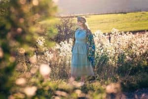 A woman in a long, light blue dress stands in a field of white flowers with green grass. She wears a headband and a colorful shawl. The background is lit with the soft, golden light of the setting sun, creating a serene and dreamy atmosphere.