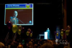 A man stands at a podium on a stage, giving a speech. A screen behind him displays the name "David Whaley" along with his title and a photo. Colorful balloons are in the foreground, and audience members are seated, watching the presentation.