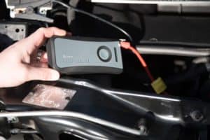 A person holds a Cobra JumpPack XL portable jump starter, showing it connected with a red cable to a vehicle's battery under the hood. The device is black and gray with a circular button. The background features car engine components.