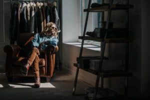 Jake Fletcher, Paul Weller's guitarist in sunglasses sits relaxed in a brown armchair, bathed in sunlight from a nearby window. They are wearing a denim shirt and brown pants. Behind them is a clothes rack with various jackets and next to them is a shelf with shoes and boxes.